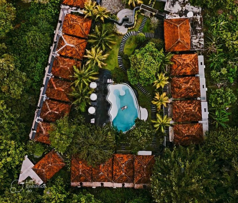 Vista aérea de um resort com prédios de telhado vermelho, vegetação exuberante e uma piscina central cercada por palmeiras.