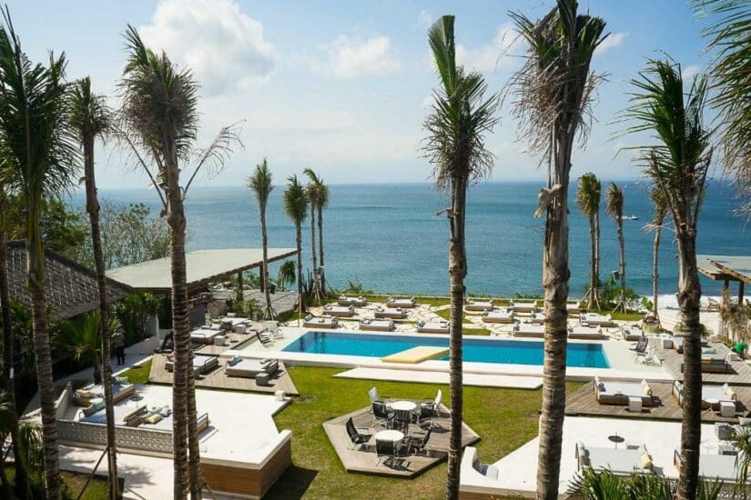 Um resort à beira-mar com piscina Onde ficar em Uluwatu, palmeiras, espreguiçadeiras e vista para o mar sob um céu parcialmente nublado.