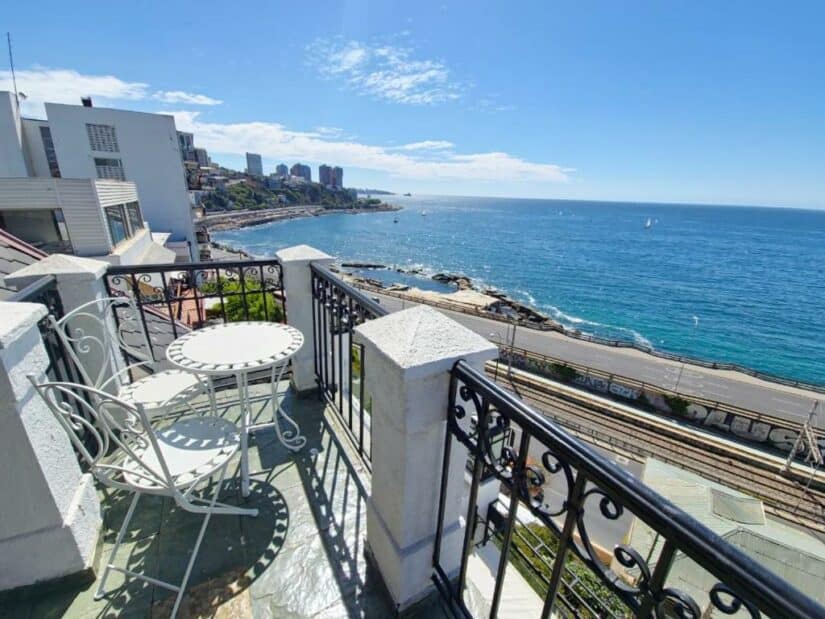 Uma pequena varanda Onde ficar em Viña del Mar com uma mesa e cadeiras tem vista para o mar, o litoral e os prédios distantes da cidade em um dia ensolarado.