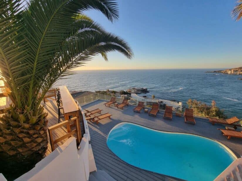 Uma piscina curva com espreguiçadeiras tem vista para o oceano ao pôr do sol, emoldurada por uma palmeira.