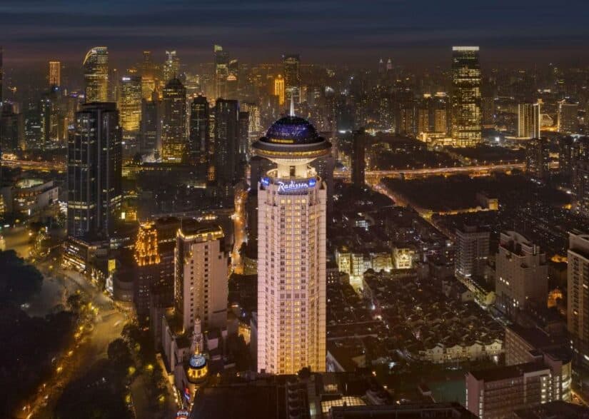 Um hotel alto e bem iluminado onde ficar em Xangai se destaca no horizonte de uma cidade à noite, cercado por edifícios iluminados.
