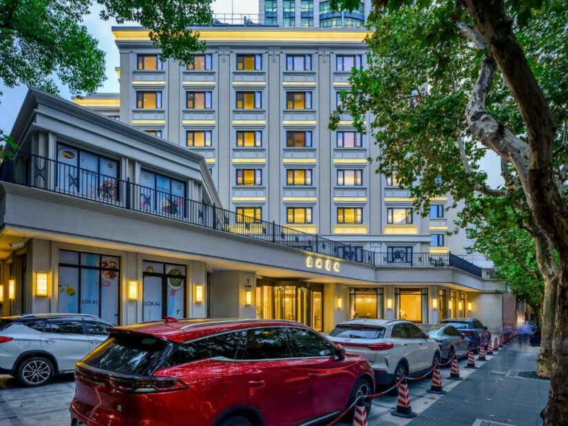 Um hotel moderno onde ficar em Xangai com janelas com iluminação quente, carros estacionados e árvores na calçada em frente.