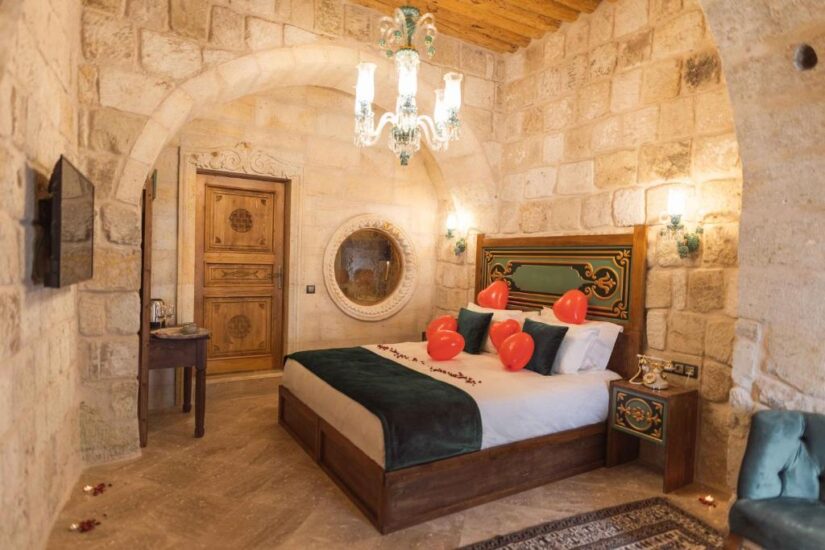 onde ficar na Capadócia Quarto com paredes de pedra, cama de casal, balões vermelhos em forma de coração, lustre e móveis de madeira ornamentados em Nevsehir na Turquia.