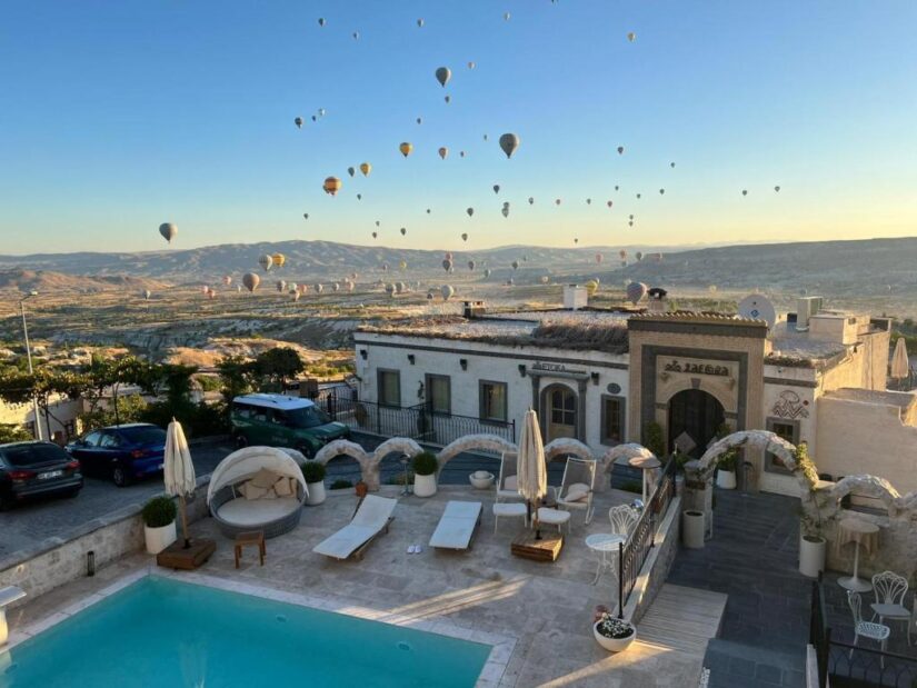 hotéis em Üçhisar na Turquia Balões de ar quente sobem sobre um vale pitoresco, com um pátio à beira da piscina e prédios brancos em primeiro plano onde ficar na Capadócia, ao nascer do sol.