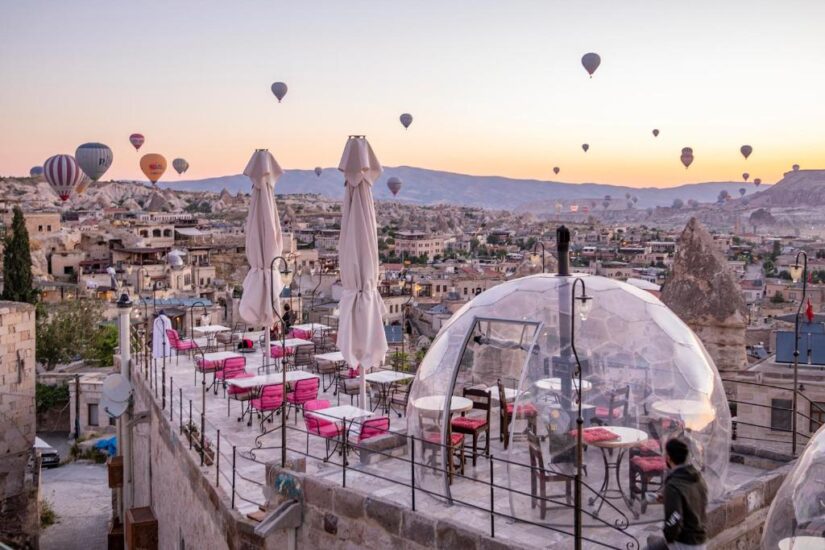 hotéis em Göreme na Turquia Café na cobertura com cadeiras cor-de-rosa, cúpula transparente e balões de ar quente sobre uma cidade histórica onde ficar na Capadócia ao nascer do sol.