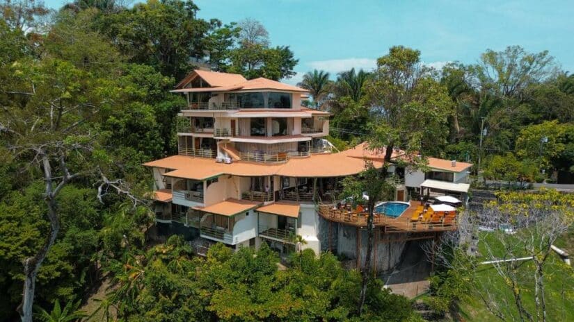 Uma villa de vários andares com varandas e uma piscina, cercada por uma vegetação exuberante e árvores tropicais.