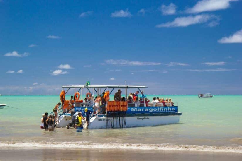 Os turistas se reúnem em torno de um barco chamado Maragolfinho em águas tropicais rasas e azul-turquesa sob um céu azul mostrando onde ficar na Praia dos Carneiros.