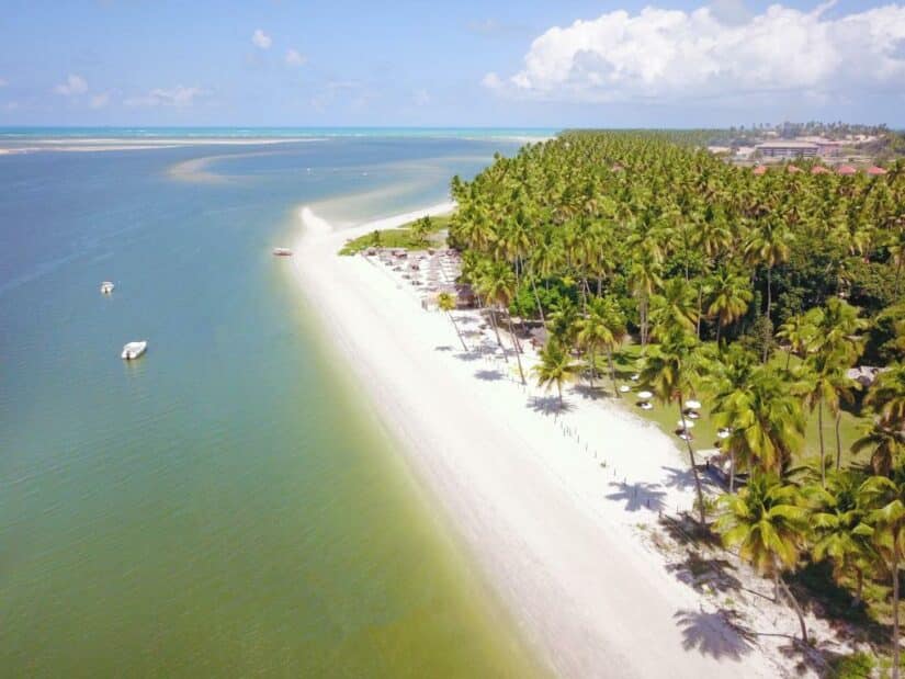 Vista aérea de uma praia tropical com areia branca, palmeiras e barcos em uma água calma azul-esverdeada, perfeita para onde ficar na Praia dos Carneiros.