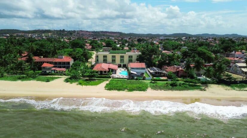 Vista aérea de casas e edifícios à beira-mar com ondas em uma costa arenosa, em uma paisagem verde.