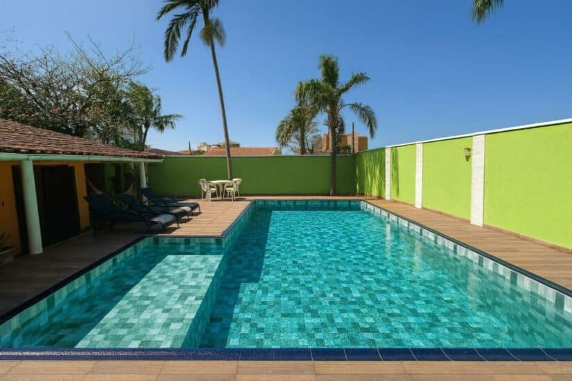 Piscina externa com água azul clara, cercada por palmeiras, espreguiçadeiras e uma parede verde brilhante Onde ficar na Praia Grande.
