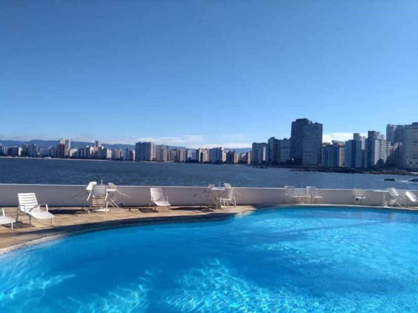 Piscina ensolarada na cobertura com espreguiçadeiras, com vista para o horizonte da cidade e para a água sob um céu azul claro.