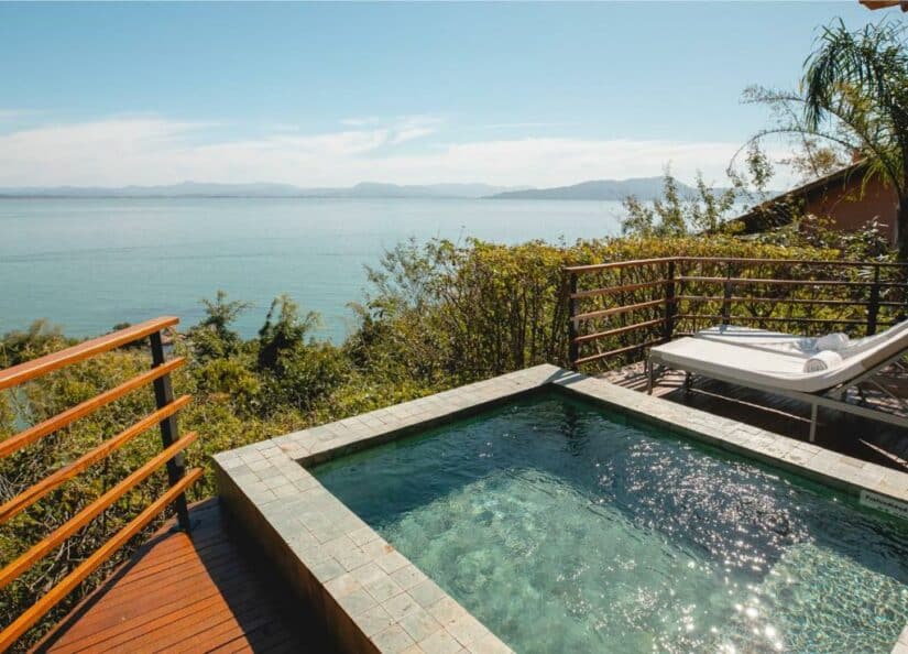 Uma pequena piscina e espreguiçadeiras têm vista panorâmica para o oceano com montanhas distantes e vegetação onde ficar em Governador Celso Ramos.
