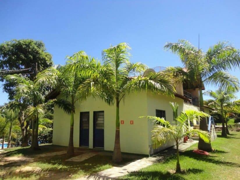 onde ficar em Holambra e Artur Nogueira Pequena casa branca com portas azuis, cercada por palmeiras e grama verde sob um céu azul claro.
