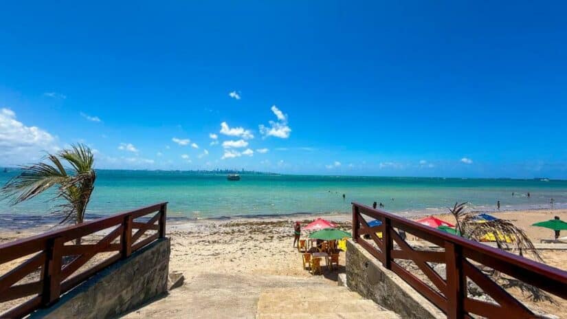 onde ficar em Itaparica na Bahia Escadas de madeira levam a uma praia de areia com guarda-sóis coloridos, mar azul calmo e um céu claro e ensolarado.