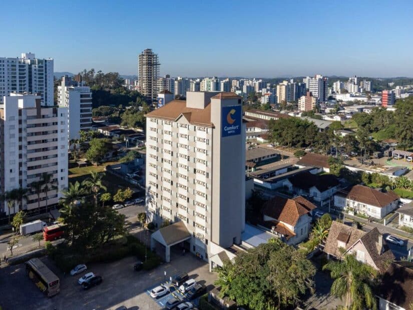 Um prédio alto de hotel onde ficar em Joinville com uma placa do Comfort Hotel em meio a prédios da cidade em um dia claro e ensolarado.