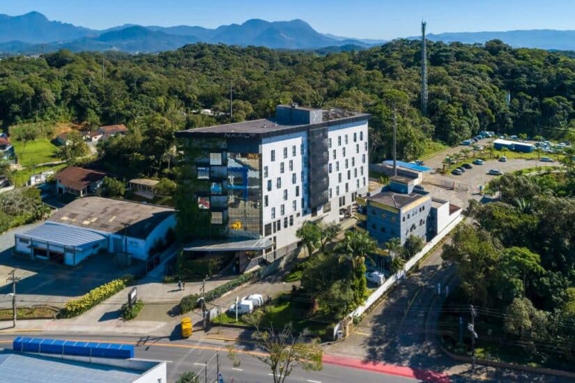 Um edifício moderno de vários andares cercado por árvores e montanhas sob um céu azul claro.