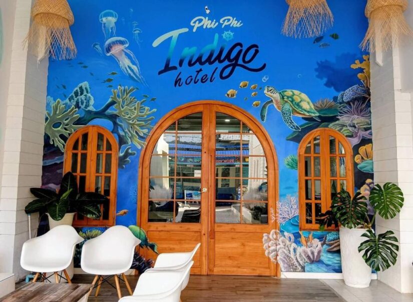 onde ficar em Koh Phi Phi na Tailândia Entrada do Phi Phi Indigo Hotel na Tailândia com um mural do oceano, portas de madeira, cadeiras brancas e vasos de plantas verdes.
