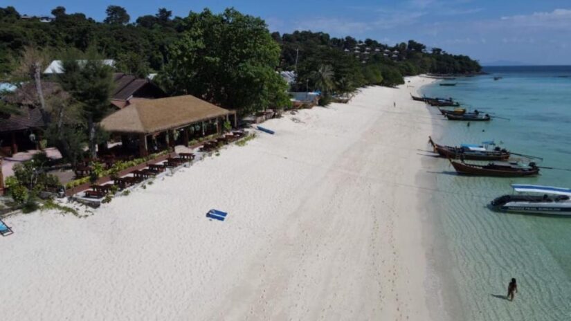 hotéis nas ilhas da Tailândia Uma praia de areia onde ficar em Koh Phi Phi com barcos em águas cristalinas, mesas ao ar livre e árvores à beira-mar sob um céu ensolarado.