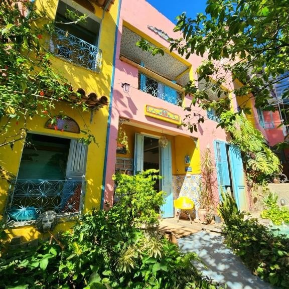 Edifício colorido com paredes amarelas e rosa, portas azuis, vasos de plantas e vegetação sob luz solar intensa onde ficar em Paquetá.