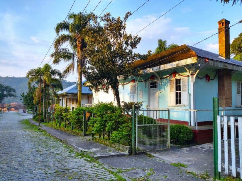 hotéis na vila de Paranapiacaba Uma charmosa casa azul-clara onde ficar em Paranapiacaba com um jardim, árvores e uma rua de paralelepípedos sob um céu ensolarado.