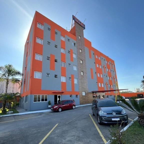 hotéis em Mauá perto de Paranapiacaba Prédio de hotel laranja e cinza de quatro andares onde ficar em Paranapiacaba com carros estacionados na frente sob um céu azul claro.