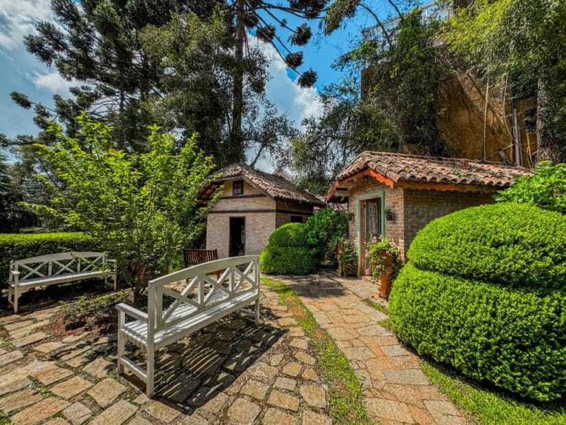hotéis em Campos do Jordão perto de São Bento do Sapucaí Dois pequenos chalés com telhados de telha onde ficar em São Bento do Sapucaí e bancos brancos em um jardim ensolarado e verdejante com caminhos de pedra.