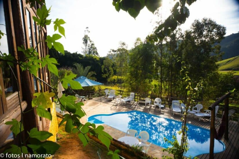 hotéis no centro de são bento do sapucaí em SP Piscina externa cercada por cadeiras brancas, deck de madeira, árvores e colinas verdes mostrando onde ficar em São Bento do Sapucaí sob luz solar intensa.