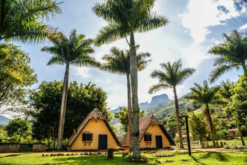 onde ficar em São Bento do Sapucaí em São Paulo Dois chalés amarelos com estrutura em A cercados por palmeiras, vegetação exuberante e montanhas distantes sob um céu azul.
