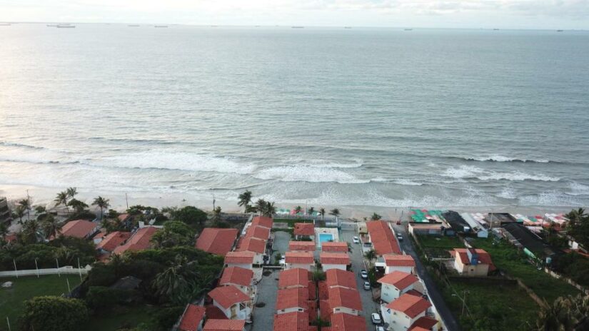 Vista aérea de casas com telhado vermelho à beira-mar com ondas suaves e navios no horizonte.