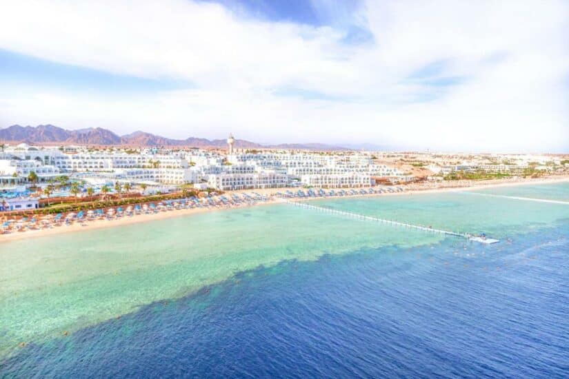 hotéis e resorts no Egito Vista aérea de um resort onde ficar em Sharm el Sheikh de praia com edifícios brancos, água azul-turquesa clara e um longo píer que se estende até o mar.