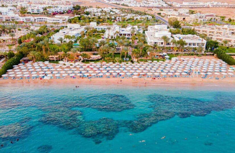 hotéis na Naama Bay no Egito Vista aérea de uma praia repleta de guarda-sóis e espreguiçadeiras, água azul clara e resorts onde ficar em Sharm el Sheikh ao fundo.