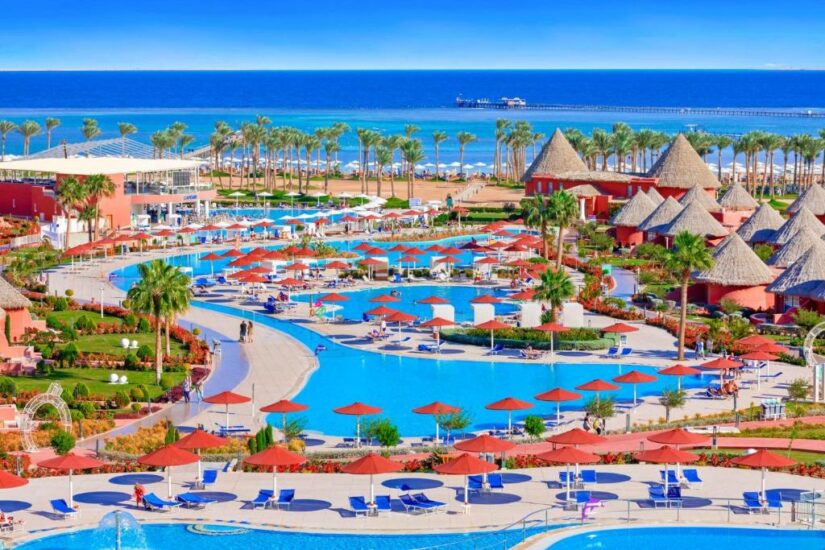 onde ficar em Sharm el Sheikh no Egito Um resort com piscinas, guarda-sóis vermelhos, palmeiras e edifícios com telhado de palha à beira-mar sob um céu azul claro.