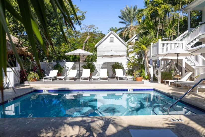 Uma serena piscina externa com espreguiçadeiras, guarda-sóis e vegetação exuberante ao lado de edifícios brancos.