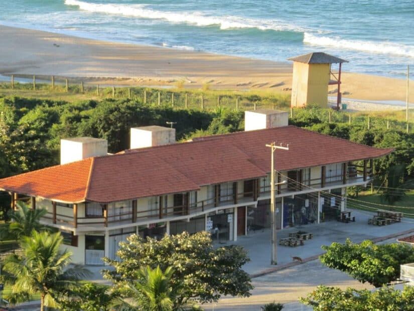 onde ficar na Guarda do Embaú ou em Gamboa em SC Um prédio de dois andares com telhado vermelho perto da praia, com ondas e uma torre de salva-vidas ao fundo.