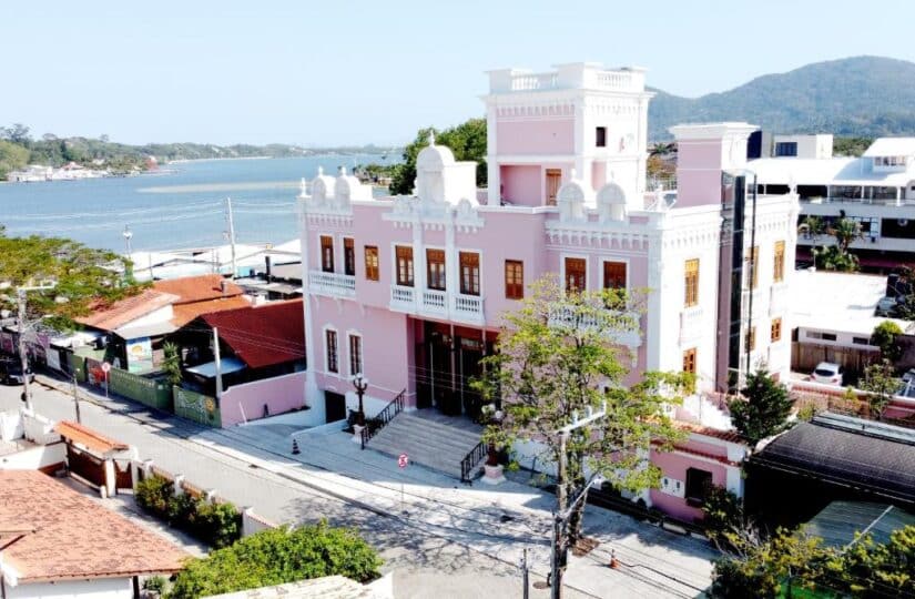 Onde ficar no Campeche e na Lagoa da Conceição em Floripa Um grande edifício histórico rosa com vista para a orla, cercado por árvores e casas próximas.