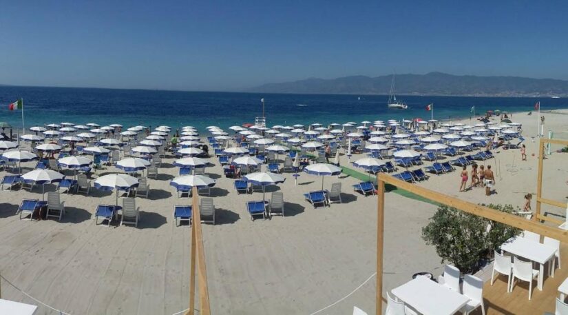 onde ficar em Reggio Calabria na Itália Fileiras de espreguiçadeiras azuis e guarda-sóis brancos em uma praia de areia à beira-mar, com montanhas à distância.