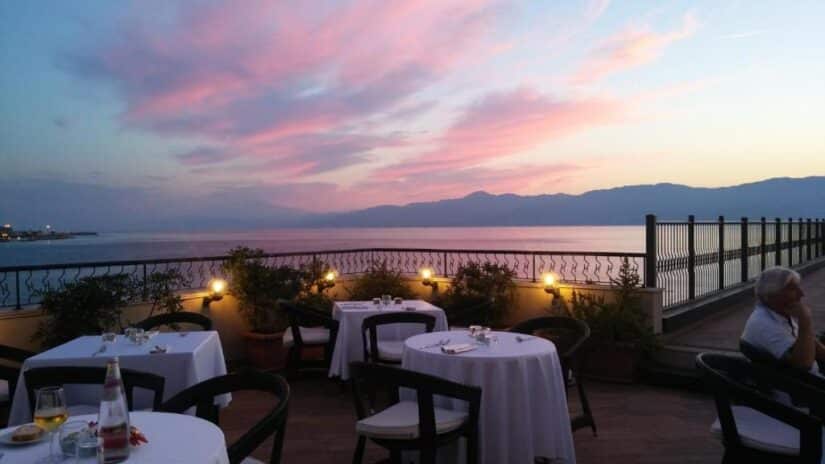 hotéis em Lungomare em Reggio Calabria na Itália Terraço onde ficar em Reggio Calabria do restaurante ao ar livre com mesas vazias, com vista para o mar ao pôr do sol com nuvens cor-de-rosa e montanhas.