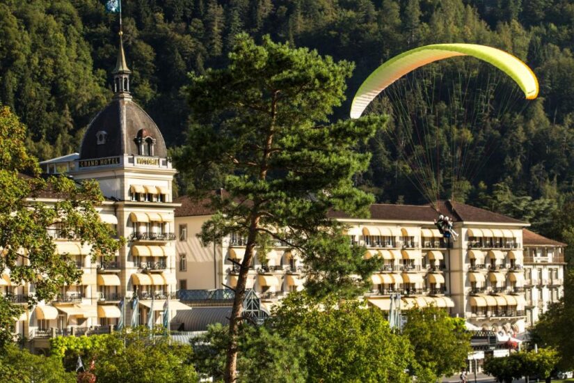 hotéis no centro de Interlaken na Suíça Um parapente sobrevoa um grande hotel Onde ficar em Interlaken com muitas varandas, cercado por árvores verdes exuberantes e colinas arborizadas.