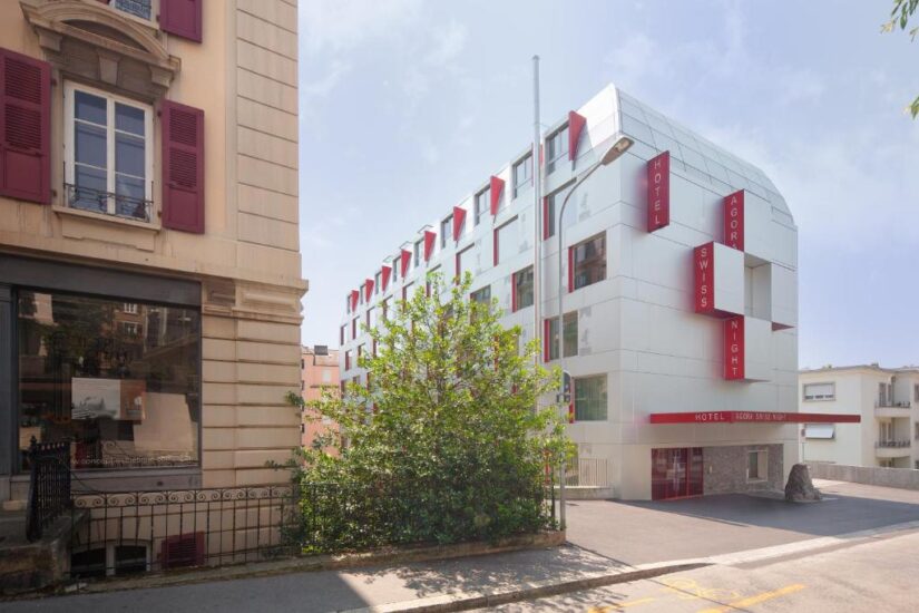 hotéis em Sous-Gare em Lausanne na Suíça Moderno edifício de hotel branco Onde ficar em Lausanne com placas vermelhas onde se lê Hotel Swiss Night ao lado de edifícios residenciais mais antigos.