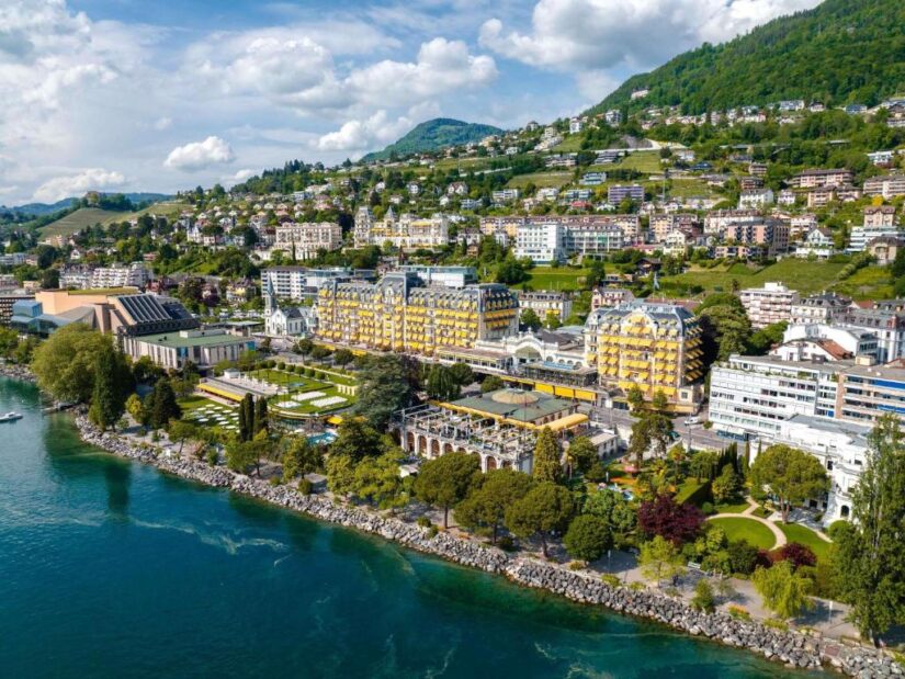 hotéis no centro de montreux na suiça Vista aérea de uma cidade à beira de um lago com prédios coloridos onde ficar em Montreux, vegetação exuberante e montanhas ao fundo.