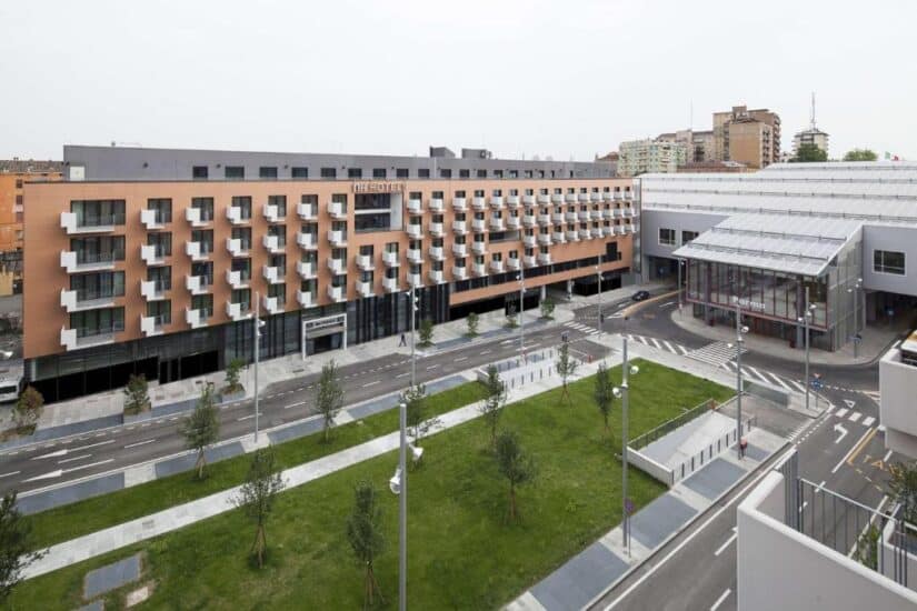hotéis perto da estação de trem de Parma na Itália Um hotel moderno onde ficar em Parma com muitas varandas fica de frente para um parque verde e uma rua vazia em uma área urbana.