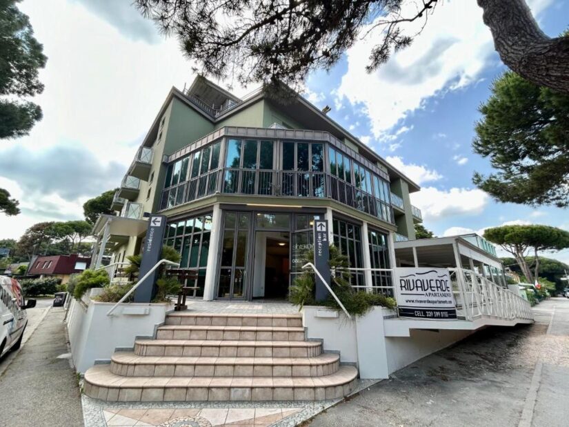 onde ficar em Ravenna na Itália Edifício moderno de três andares com grandes janelas de vidro, cercado por árvores, com uma rampa e escadas na entrada.